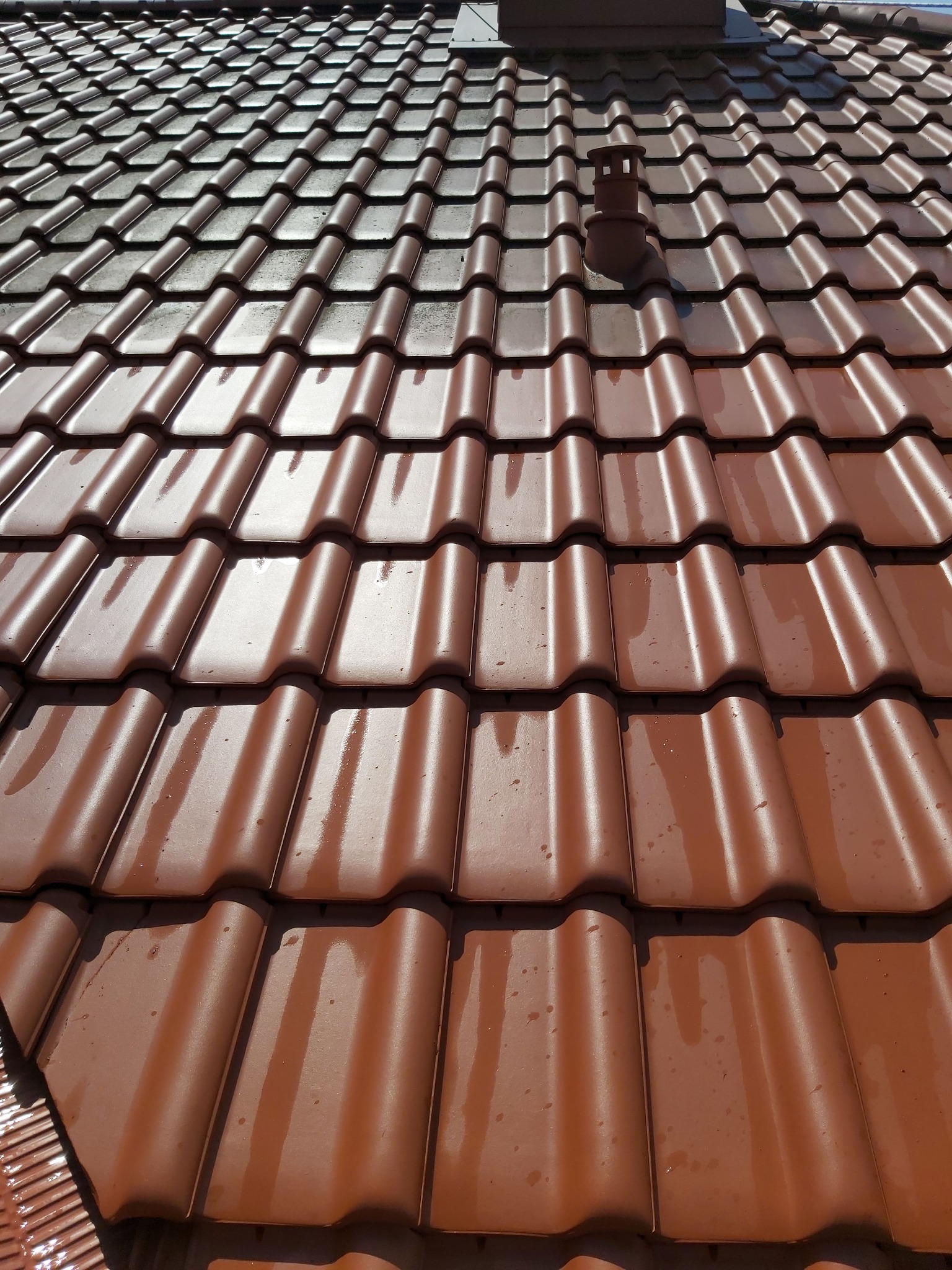 Błyszcząca, brązowa dachówka ceramiczna widziana pod kątem, z widocznym kominem wentylacyjnym i kroplami wody po deszczu lub myciu.