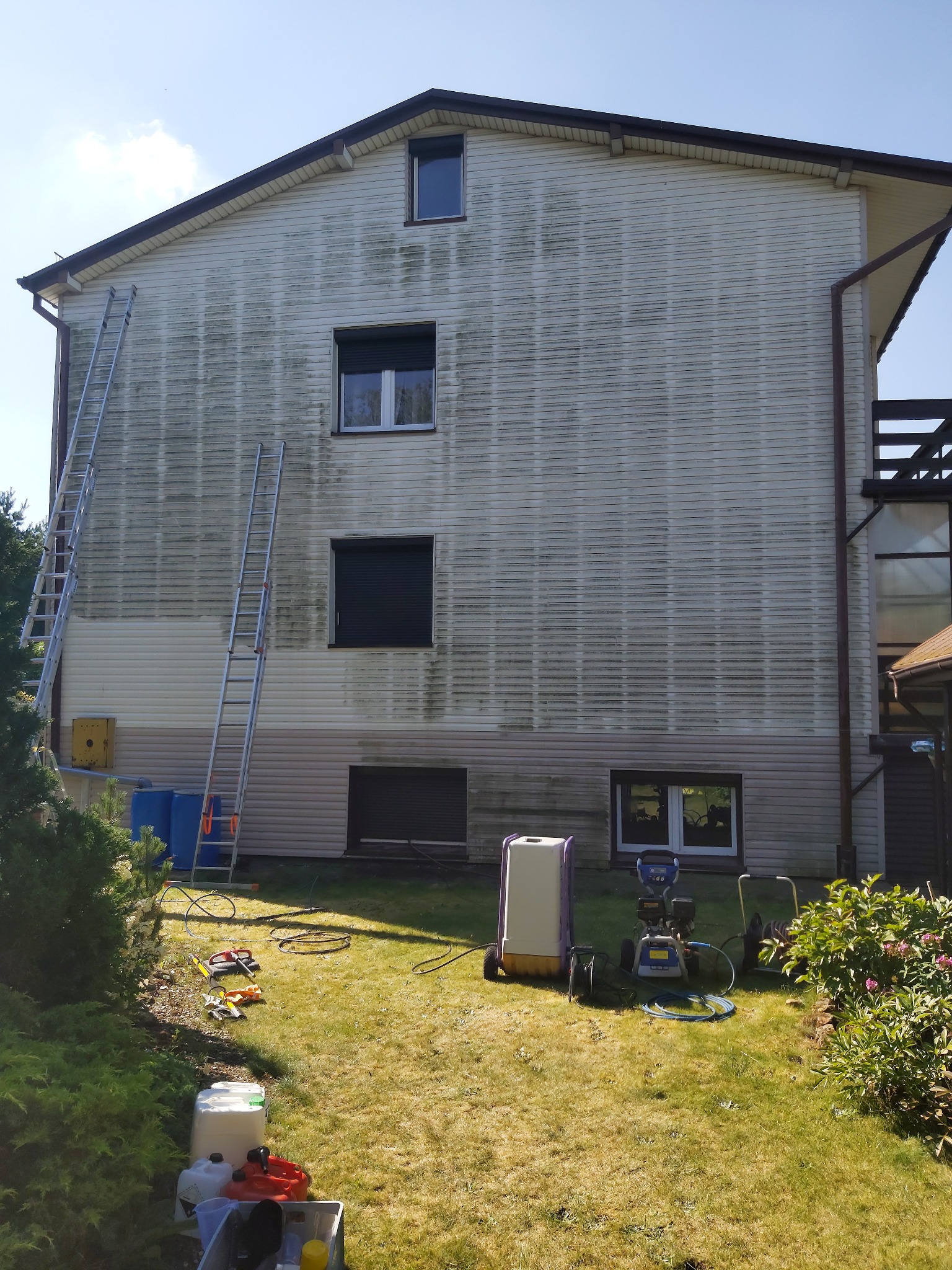 Boczna ściana domu z sidingiem, częściowo oczyszczona z zabrudzeń, dwie drabiny opierają się o ścianę, na trawniku rozłożone narzędzia i sprzęt do czyszczenia.