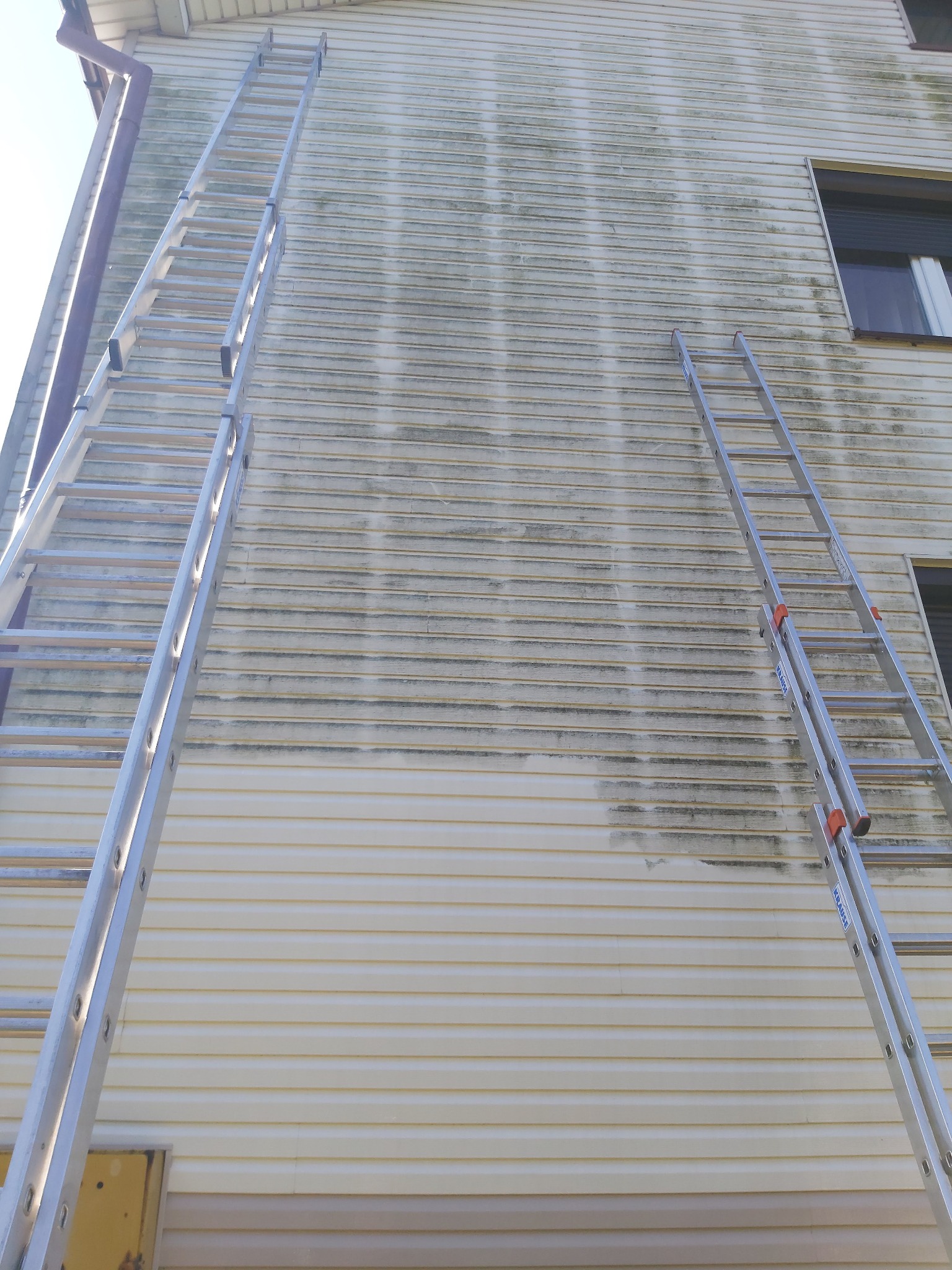 Dwie aluminiowe drabiny opierają się o elewację budynku z sidingu, widoczne ślady czyszczenia i pozostałości brudu, okna z roletami w tle.