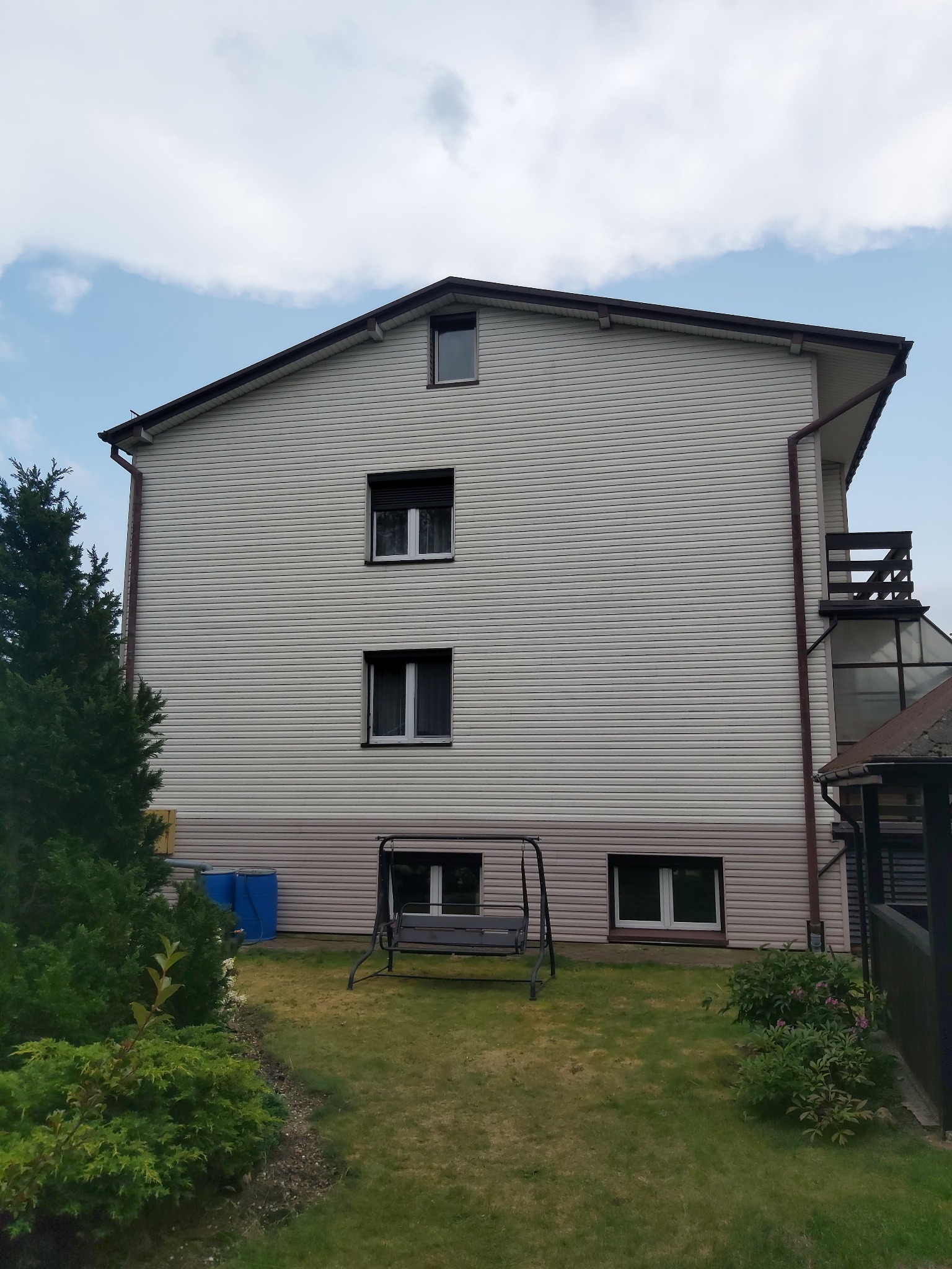 Boczna ściana dwupiętrowego domu z sidingiem w odcieniach beżu, widoczne okna z roletami, brązowe rynny, mały balkon i huśtawka ogrodowa na trawniku.