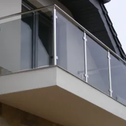 Nowoczesna, szklana balustrada balkonowa z elementami metalowymi, zamocowana na jasnym, otynkowanym balkonie. Widok z dołu, fragment dachu w tle.