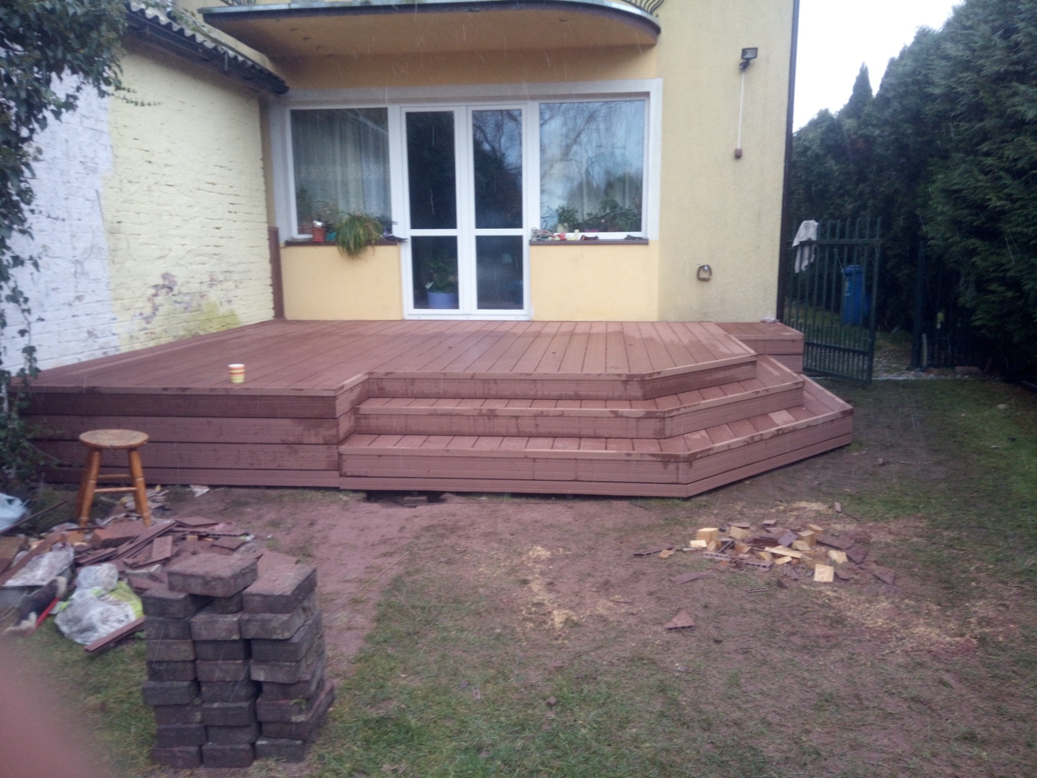 Nowo wybudowany taras z brązowych desek kompozytowych z kilkoma schodkami, prowadzący do białych drzwi balkonowych w żółtym domu, widoczne pozostałości po budowie: sterta kostki brukowej...