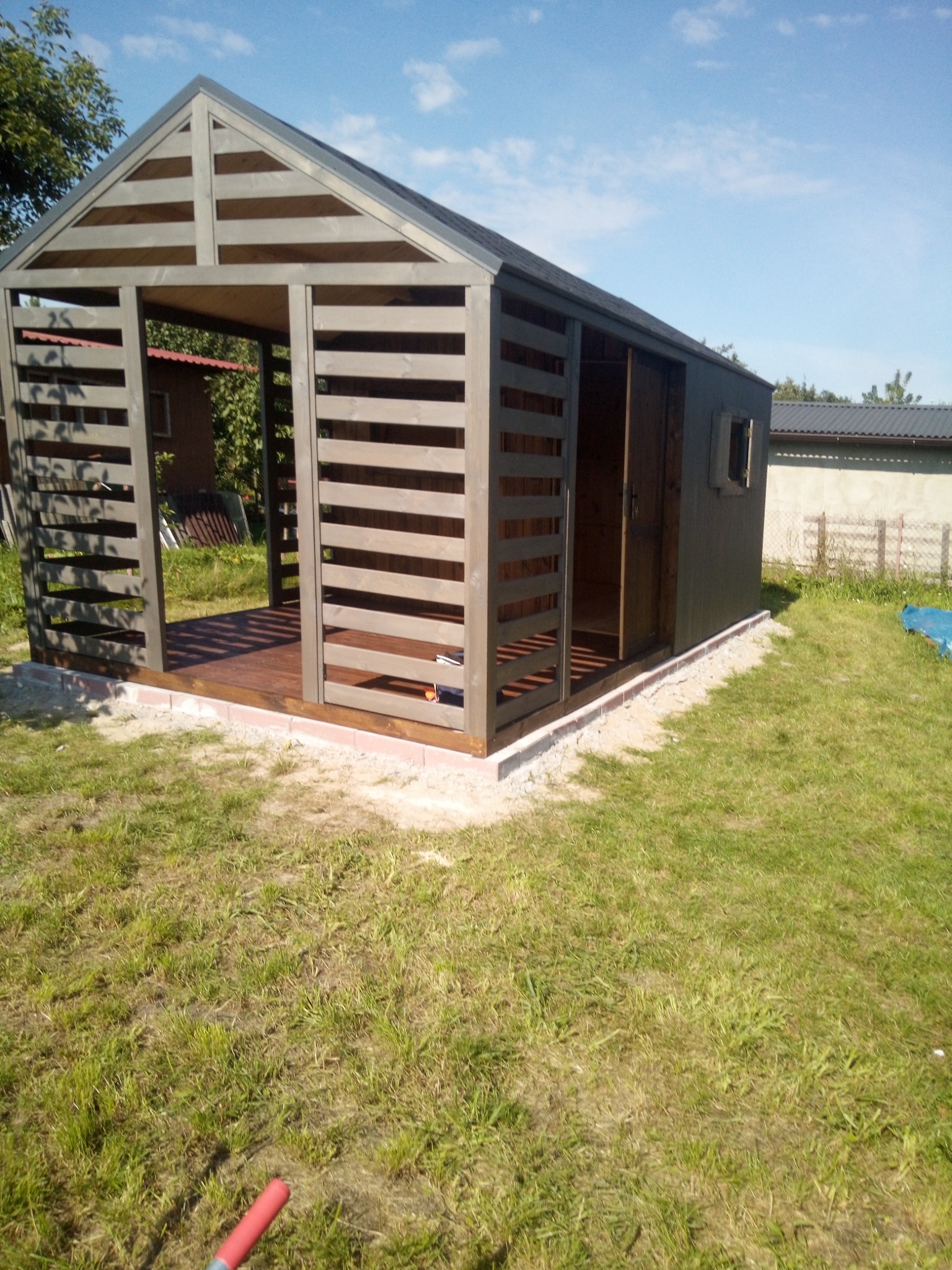 Nowoczesna, szara konstrukcja z drewna z ażurowymi ścianami i podłogą z desek, prawdopodobnie altana lub domek narzędziowy, na trawniku w słoneczny dzień.