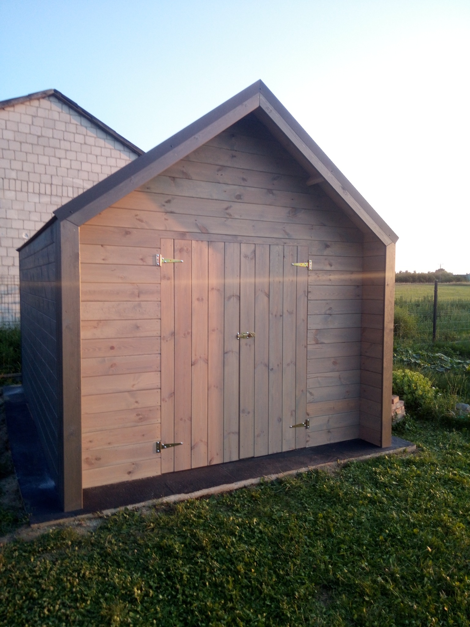 Mała, drewniana konstrukcja z dwuspadowym dachem, prawdopodobnie domek narzędziowy lub schowek ogrodowy, z podwójnymi drzwiami zabezpieczonymi na złoty zatrzask, ustawiona na trawniku przed...