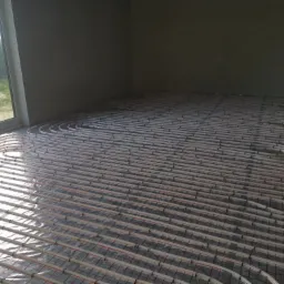 Rozłożone rury ogrzewania podłogowego na folii izolacyjnej w surowym wnętrzu z dużym oknem.