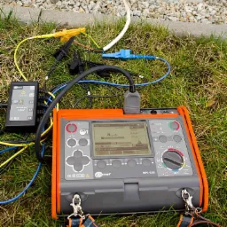 Miernik MPI-530 firmy Sonel z podłączonymi przewodami pomiarowymi na trawie w Olsztynie, widoczny ekran urządzenia z wynikami pomiarów.