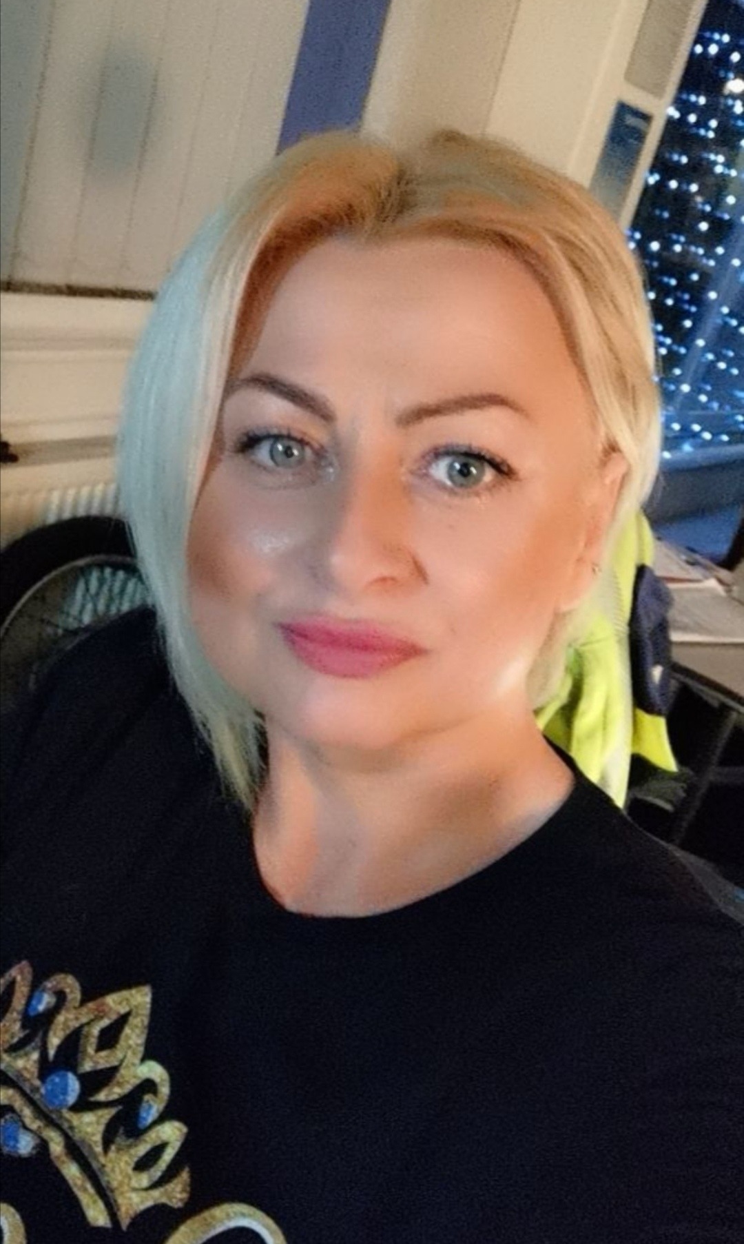 Portret kobiety o blond włosach i niebieskich oczach, ubranej w czarną koszulkę z motywem korony, w tle widoczne elementy wnętrza i dekoracje.