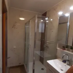 Nowoczesna łazienka po remoncie: kabina prysznicowa z chromowanymi elementami, umywalka z szafką, marmurowe płytki na ścianach, jasne oświetlenie sufitowe i zabezpieczenie podłogi kartonem.