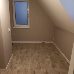 Usługi Budowlane MatBud Mateusz Malec - Poddasze po remoncie z drewnopodobną podłogą z płytek, beżowe ściany i białe listwy przypodłogowe, widoczne okno z białymi ramami.
