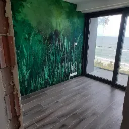 Pokój z ułożoną podłogą z paneli imitujących drewno, ściana z fototapetą z motywem trawy i widokiem na morze przez duże okno balkonowe, widok zza surowej ściany z cegłami.