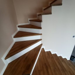 Usługi Budowlane MatBud Mateusz Malec - Drewniane schody wachlarzowe z białymi podstopnicami, widok z dołu, ściany w odcieniach beżu, częściowo widoczne narzędzia malarskie.