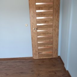 Usługi Budowlane MatBud Mateusz Malec - Zamknięte, drewniane drzwi z poziomymi, metalowymi wstawkami, zamontowane w świeżo wyremontowanym pomieszczeniu z panelami podłogowymi i gładkimi, szarymi ścianami.