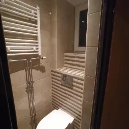 Beżowa łazienka z poziomymi pasami płytek, wisząca toaleta, grzejnik drabinkowy i okno w zabudowie.