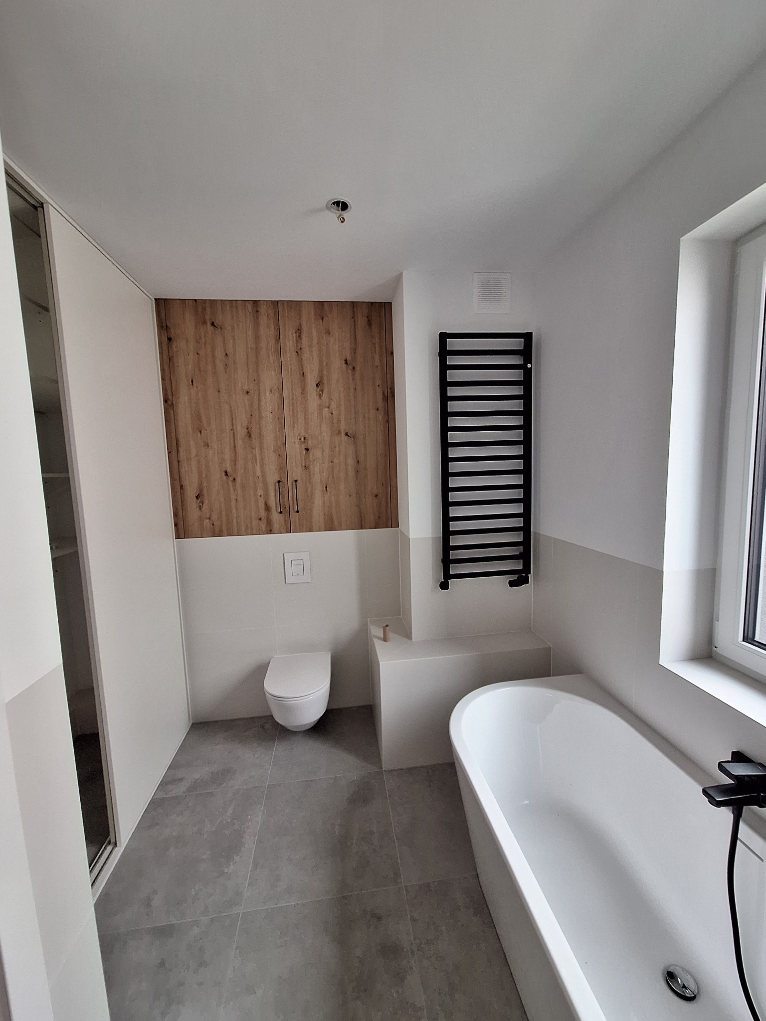 Nowoczesna łazienka z drewnianą zabudową szafek, grzejnikiem drabinkowym i białą wanną. Szare płytki podłogowe i białe ściany tworzą minimalistyczny design.