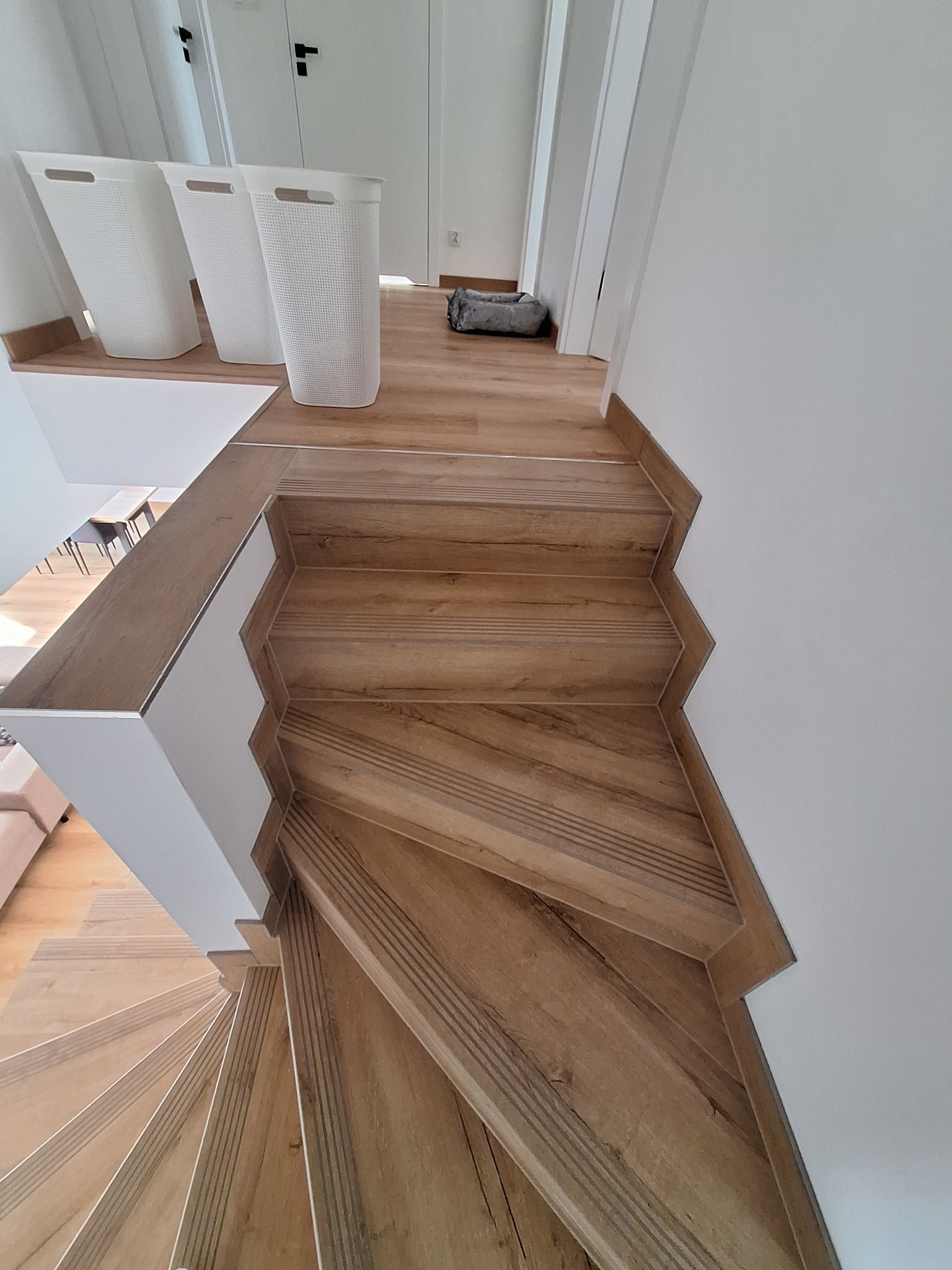 Spiralne schody wyłożone płytkami imitującymi drewno, z białymi balustradami i trzema białymi koszami na pranie na górze. Widok z góry, jasne wnętrze.