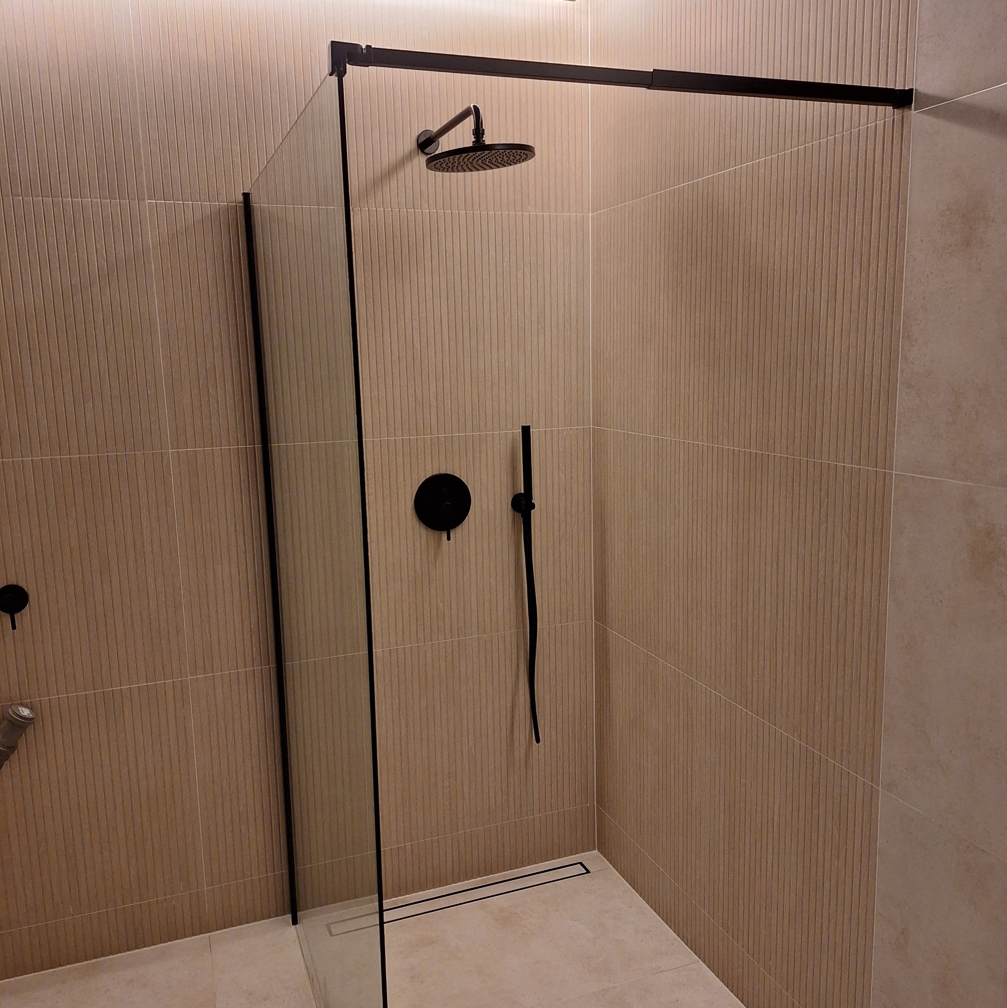 Nowoczesna kabina prysznicowa z czarną armaturą i beżowymi, pionowo żłobionymi płytkami. Minimalistyczny design z odpływem liniowym.
