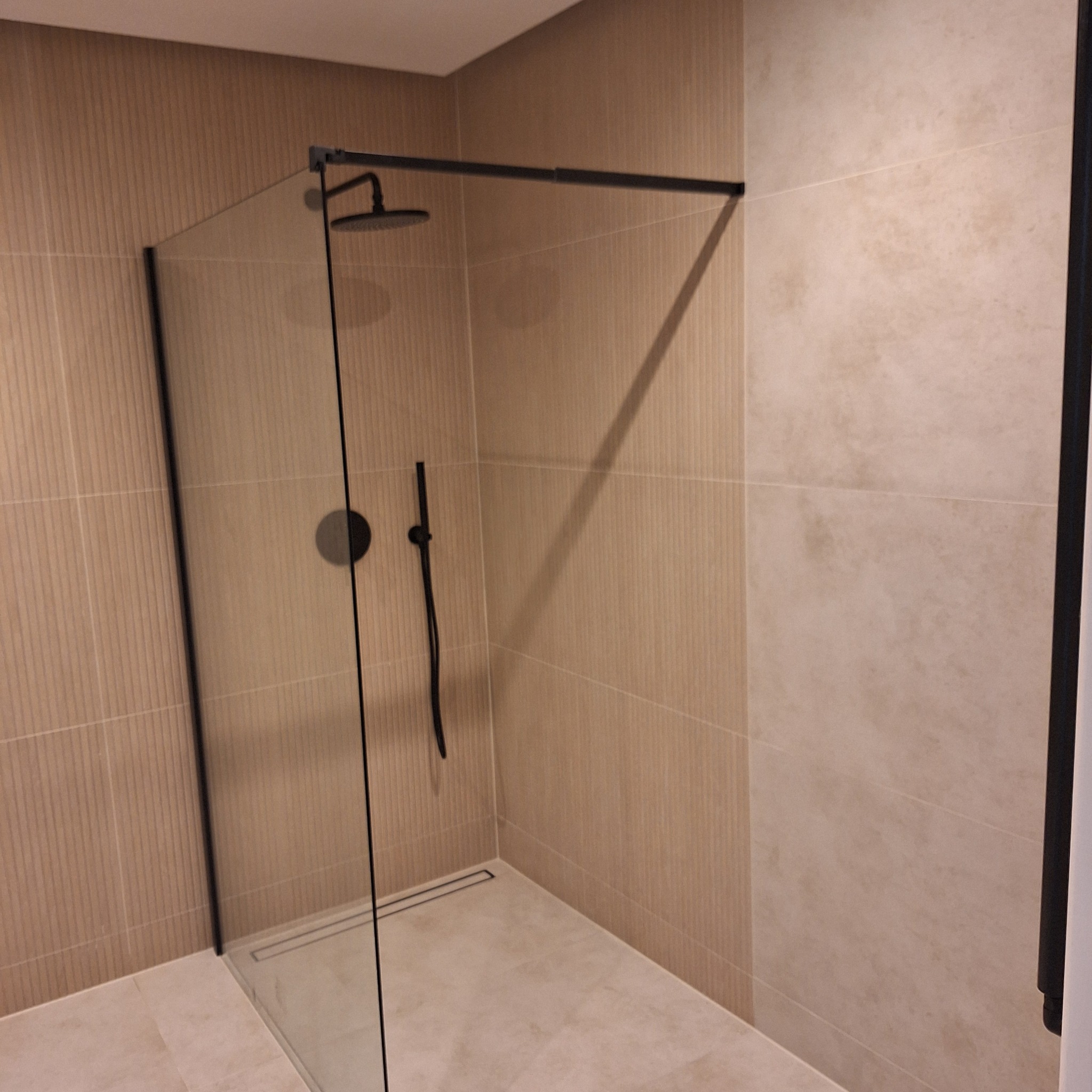 Nowoczesna kabina prysznicowa z czarnymi detalami na tle beżowych płytek. Minimalistyczny design, widoczny odpływ liniowy. Ukończona realizacja.