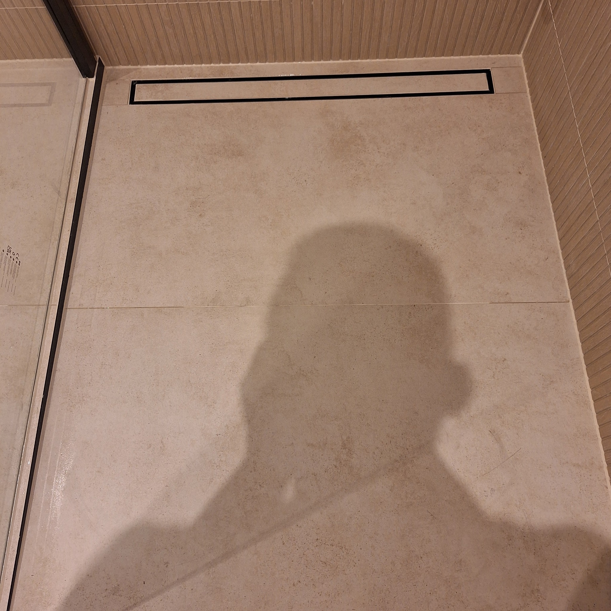 Beżowe płytki podłogowe w kabinie prysznicowej z odpływem liniowym i szklaną ścianką. Cień osoby na podłodze.