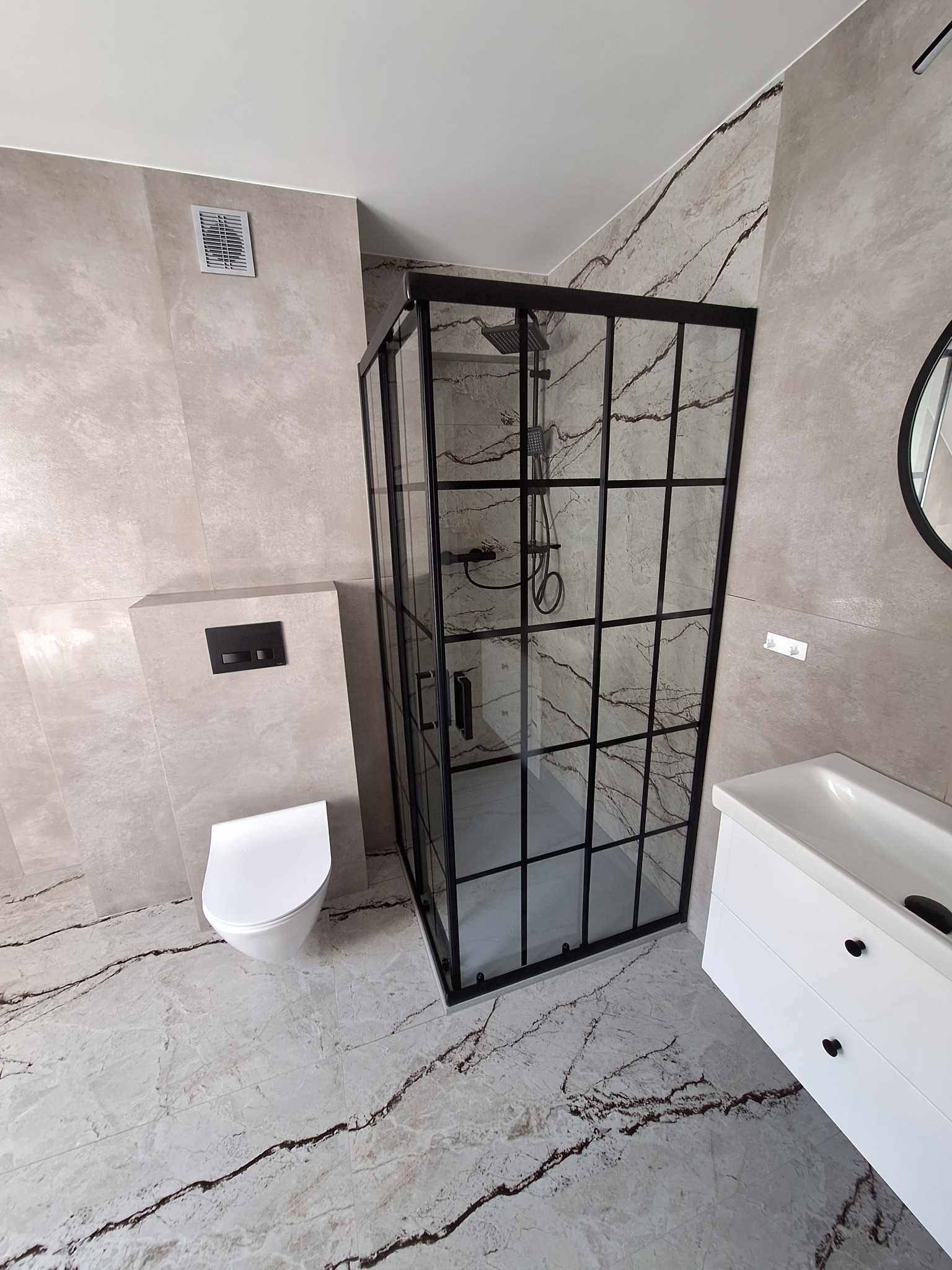 Nowoczesna łazienka z kabiną prysznicową typu walk-in z czarnymi profilami, umywalką z szafką i podwieszaną toaletą. Płytki imitujące marmur na podłodze i ścianach.