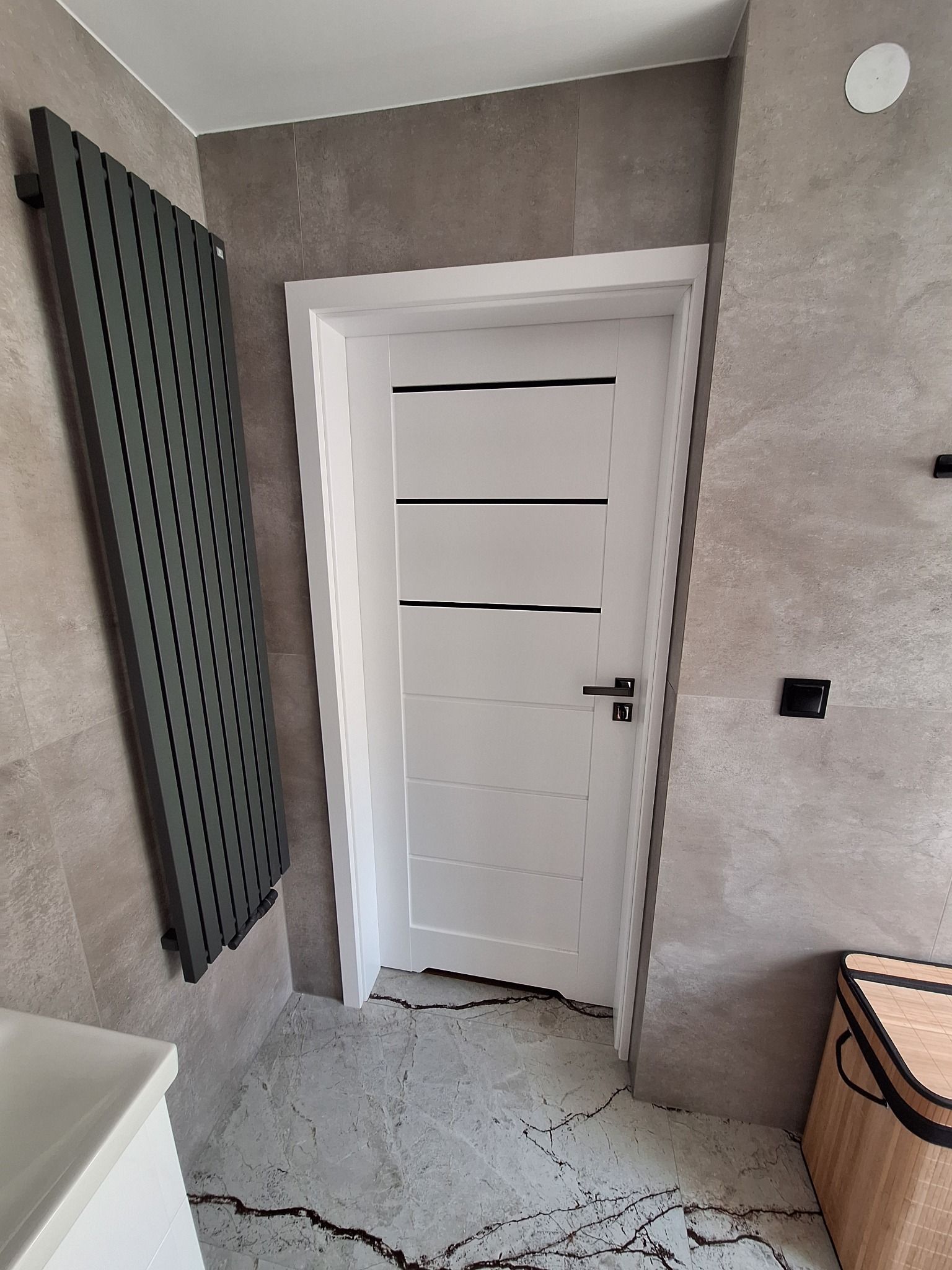 Nowoczesna łazienka z białą, minimalistyczną stolarką drzwiową z czarnymi akcentami, szarym grzejnikiem drabinkowym i płytkami imitującymi kamień.