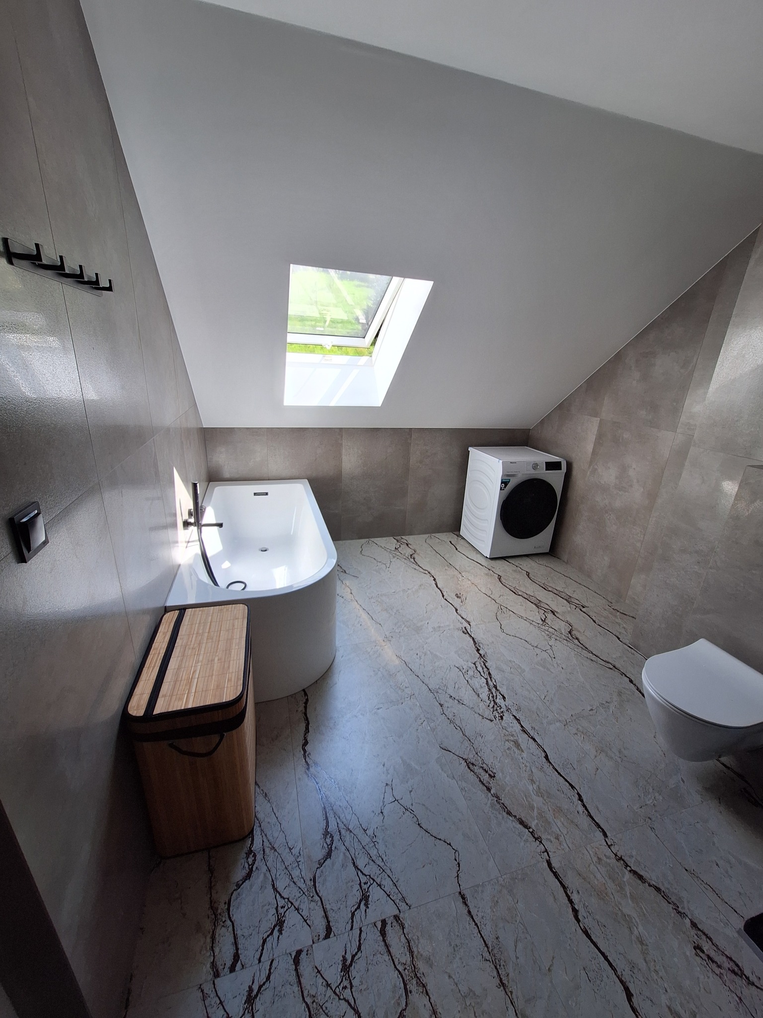 Wykończona łazienka na poddaszu z marmurową podłogą, białą wanną, pralką i oknem dachowym. Szaro-beżowe płytki na ścianach. Nowoczesny design.