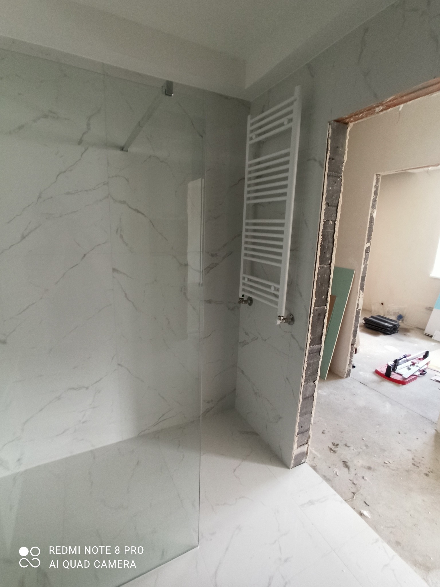 Wykończona kabina prysznicowa z białymi płytkami imitującymi marmur, grzejnik drabinkowy na ścianie, widok na surowy otwór drzwiowy i narzędzia na podłodze w trakcie remontu.