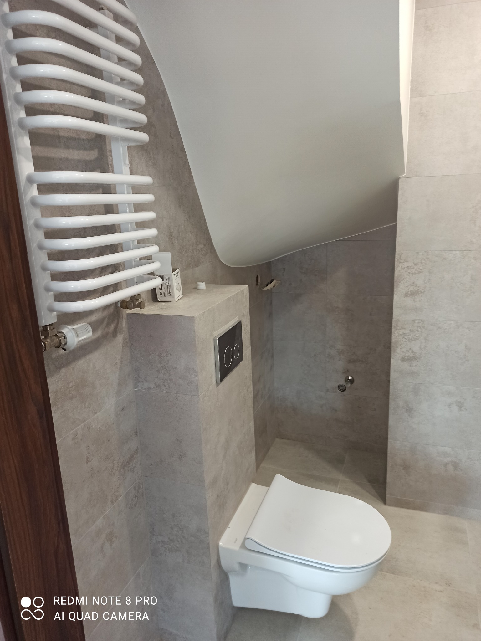 Nowoczesna łazienka z szarymi płytkami, podwieszaną toaletą z czarnym panelem sterującym, białym grzejnikiem drabinkowym i skośnym sufitem.