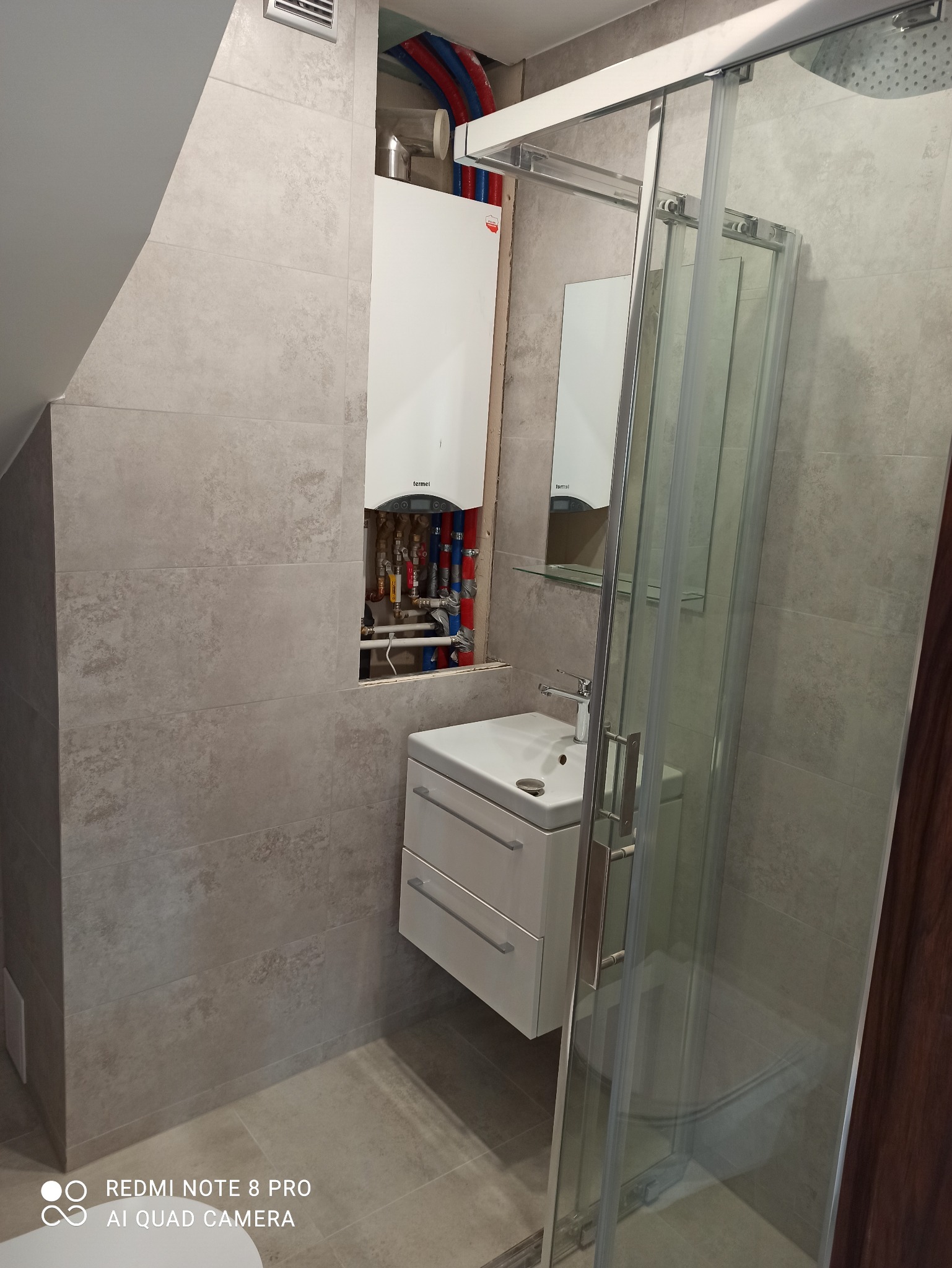 Świeżo wyremontowana łazienka z beżowymi płytkami, kabiną prysznicową z przesuwnymi drzwiami, białą szafką z umywalką i ukrytym za nią piecem gazowym z widocznymi rurami instalacji.