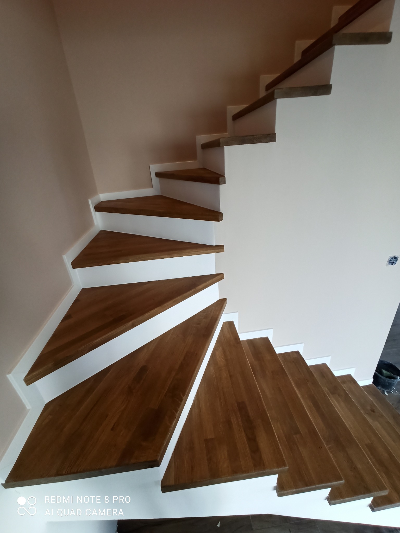 Drewniane schody wachlarzowe z białymi podstopnicami, widok z dołu, ściany w odcieniach beżu, częściowo widoczne narzędzia malarskie.