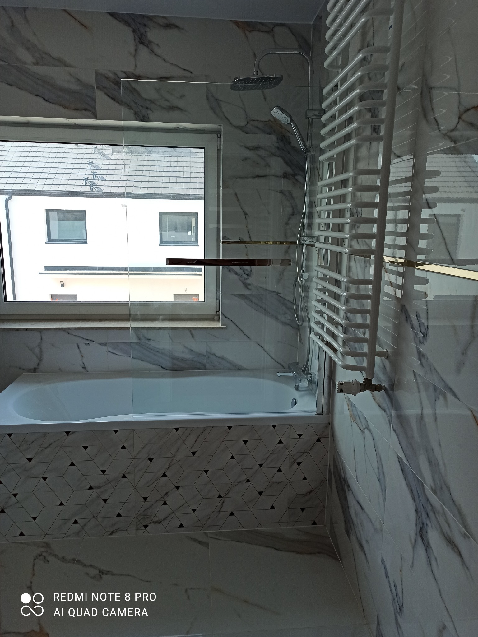 Wykończona łazienka z wanną obudowaną płytkami w geometryczne wzory, marmurowe płytki na ścianach, szklany parawan nawannowy, grzejnik drabinkowy i widok z okna na biały budynek z szarą dachówką.