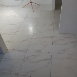 Wyłożona jasnymi, marmurkowymi płytkami podłoga w trakcie prac wykończeniowych, widoczna lampa budowlana na trójnogu i gniazdka elektryczne w ścianie.