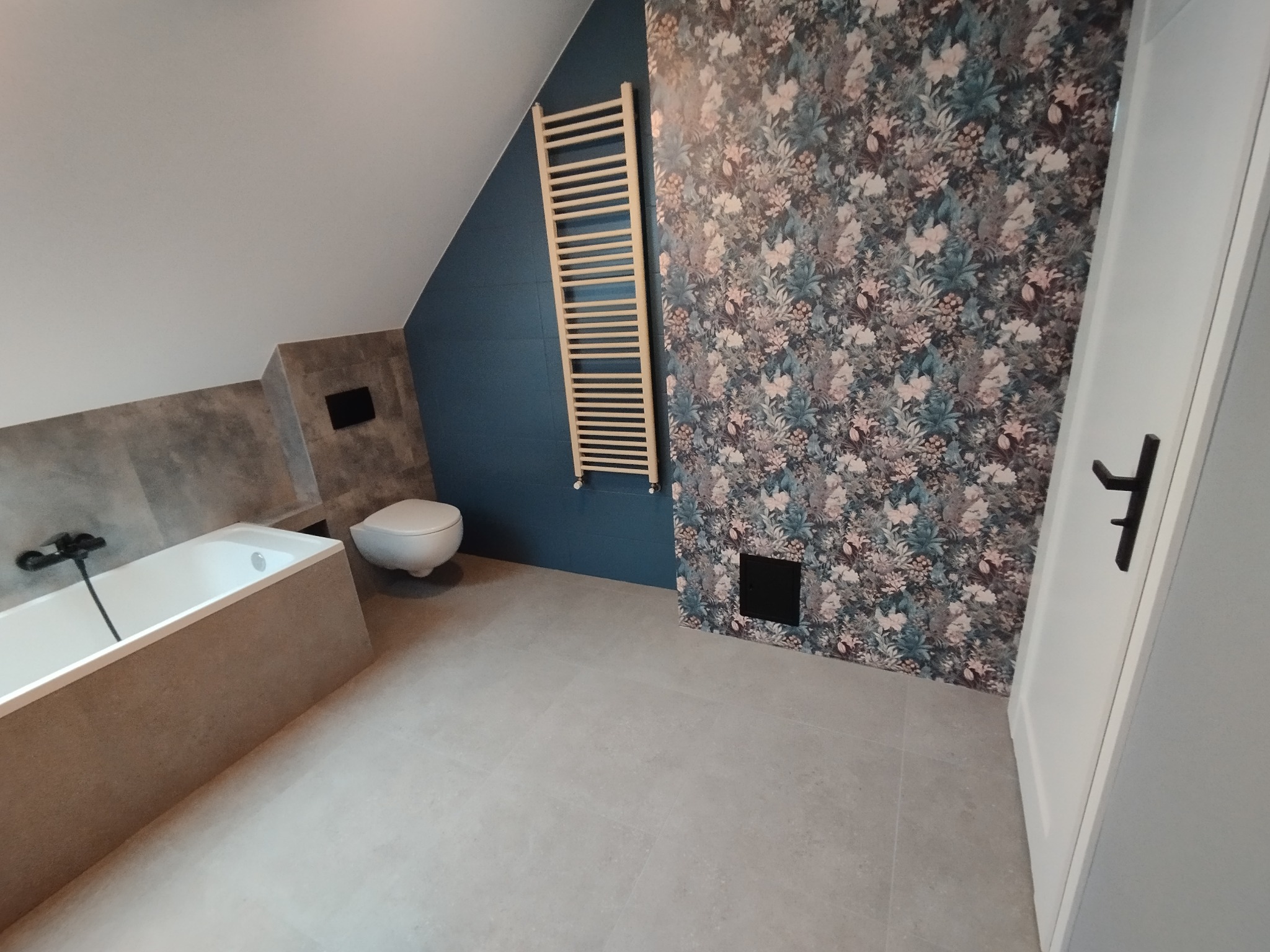 Wykończona łazienka na poddaszu ze ścianą w kwiaty, grzejnikiem drabinkowym, wanną obudowaną płytkami i szarą podłogą. Uwagę zwraca skośny sufit i nietypowe połączenie kolorów.