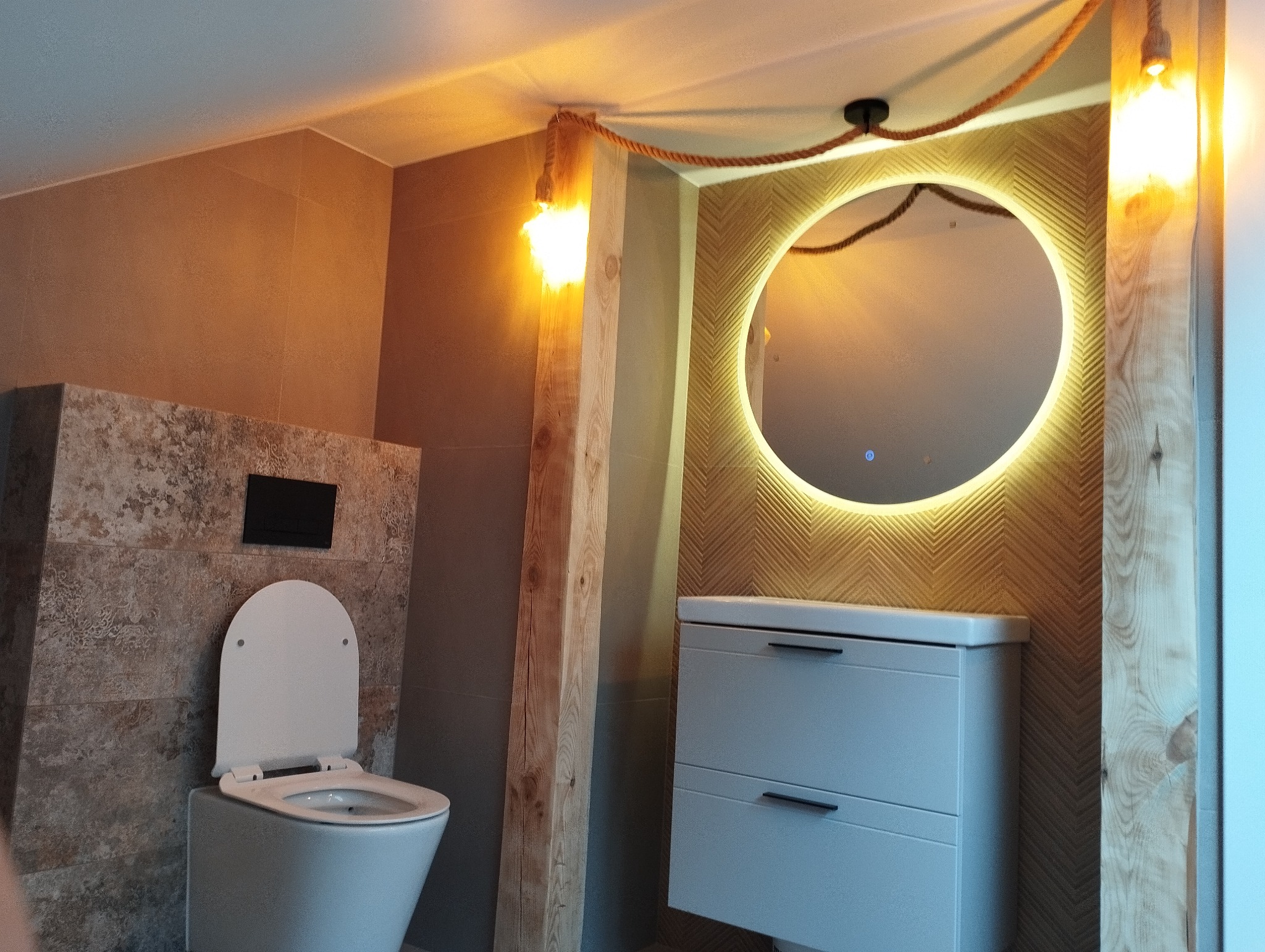 Nowoczesna łazienka z toaletą, umywalką z szafką i okrągłym lustrem z podświetleniem LED. Dekoracyjne oświetlenie z liną jutową. Ściany wykończone w różnych odcieniach brązu.