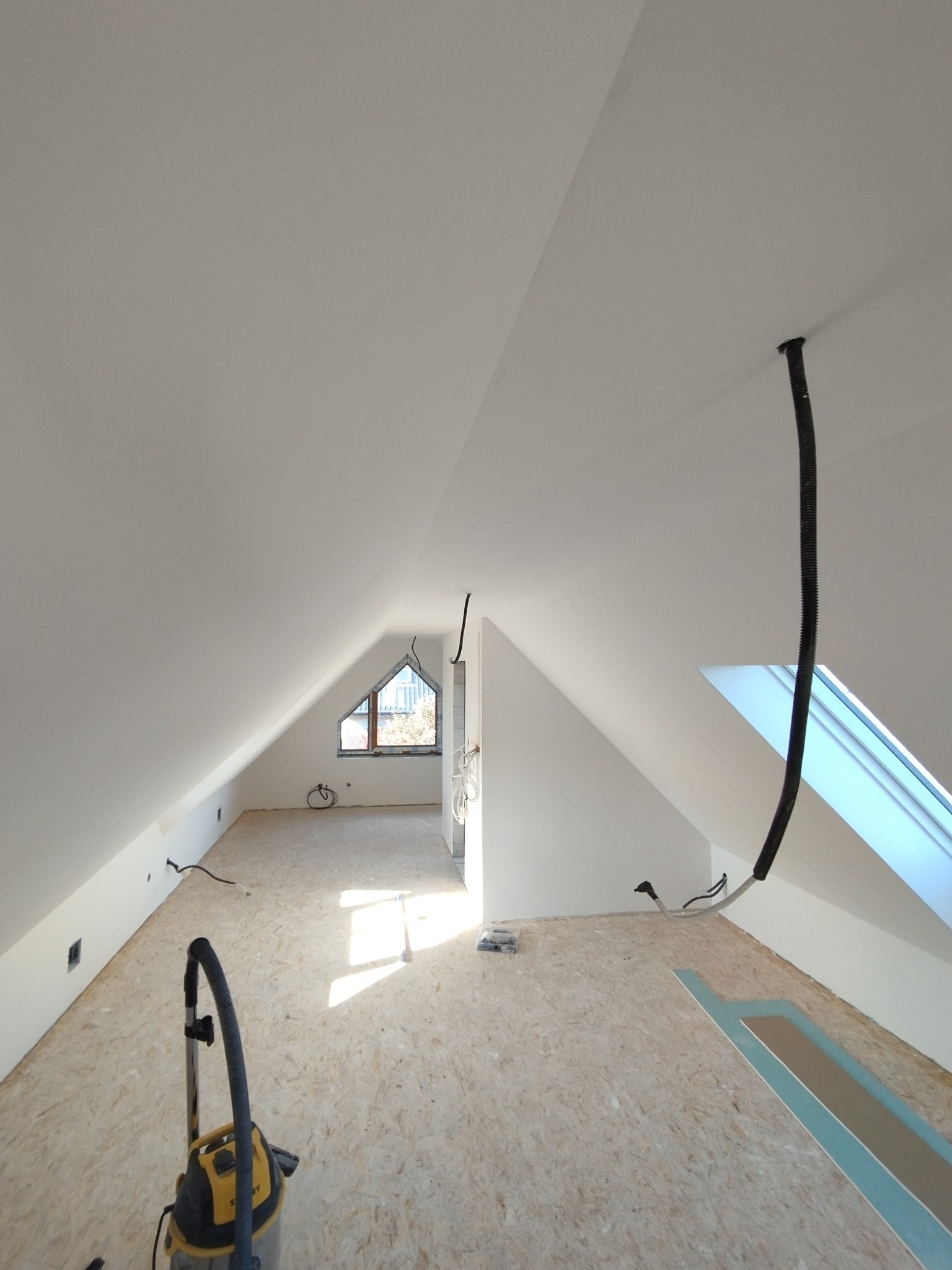 Białe poddasze ze skośnymi ścianami i oknami dachowymi w trakcie prac wykończeniowych, widoczny odkurzacz i wystające przewody.