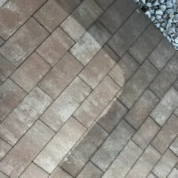 Ułożona pod kątem kostka brukowa w odcieniach brązu i szarości, sąsiadująca z obszarem wysypanym drobnymi, szarymi kamieniami. Widoczny cień częściowo zakrywa powierzchnię kostki. W górnym rogu...