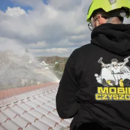 Pracownik w kasku i bluzie z logo firmy spryskuje wodą dach pokryty białą pianą z detergentu; w tle widoczne zabudowania i błękitne niebo z chmurami.