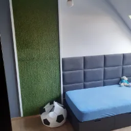 Pokój dziecięcy z łóżkiem z szarym zagłówkiem w kwadraty, niebieskim materacem i pluszowym misiem, obok ściana z panelem imitującym trawę i pufa w kształcie piłki nożnej.