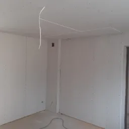 Montaż płyt kartonowo-gipsowych na ścianach i suficie w pomieszczeniu podczas remontu, widoczne kable wystające z sufitu, szpachlowane łączenia płyt.