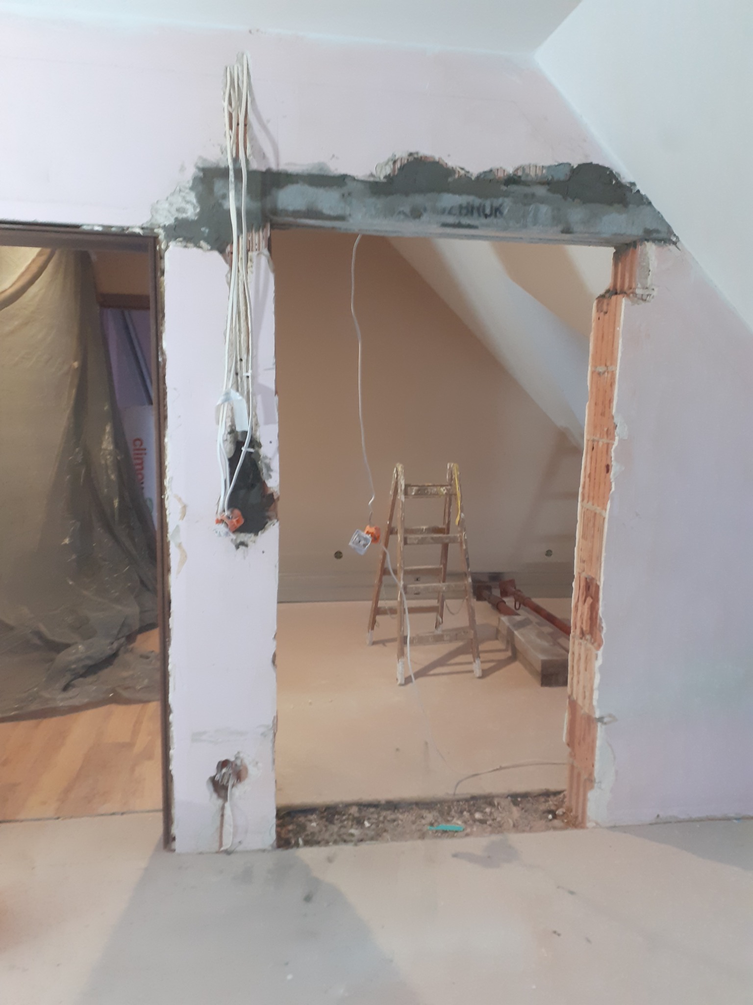 Wnętrze w trakcie prac remontowych: widoczne ślady po kuciu, wystające przewody elektryczne, fragment ściany z cegły, drabina malarska w tle, zabezpieczone folią drzwi w sąsiednim pomieszczeniu.