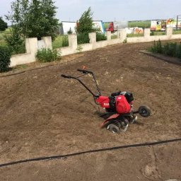 płatmar - Czerwony glebogryzarek spalinowy stoi na świeżo przekopanej ziemi, widoczny fragment nawadniania kroplowego i betonowe ogrodzenie w tle.