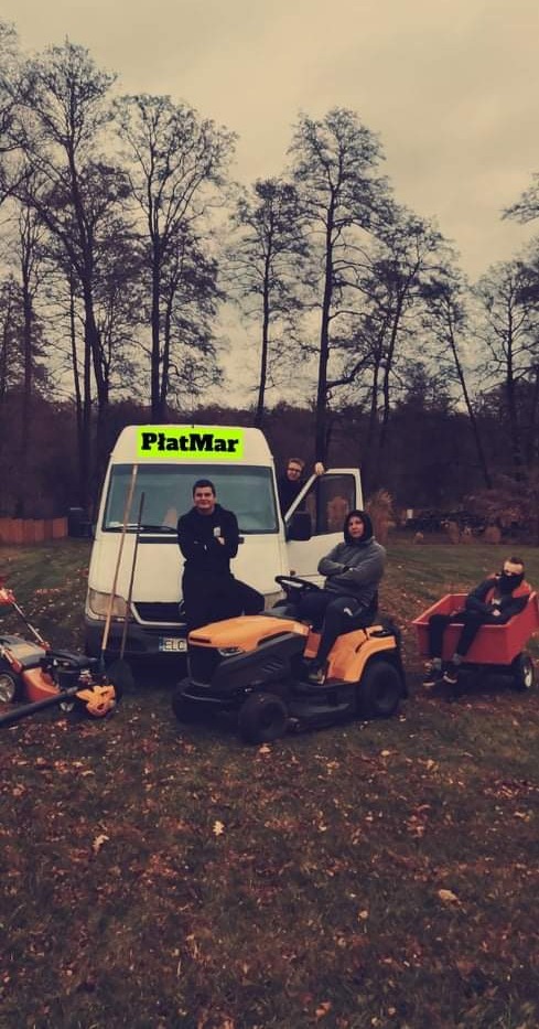 Zespół pracowników z kosiarkami i wózkiem na trawie przed białym busem z zielonym napisem PłatMar, drzewa w tle.