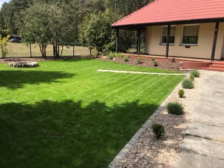 Perfekcyjnie przystrzyżony trawnik z widocznymi pasami po kosiarce, obrzeża z kostki brukowej i kamyków, z tyłu domu widoczna strefa ogniska z kamieni.