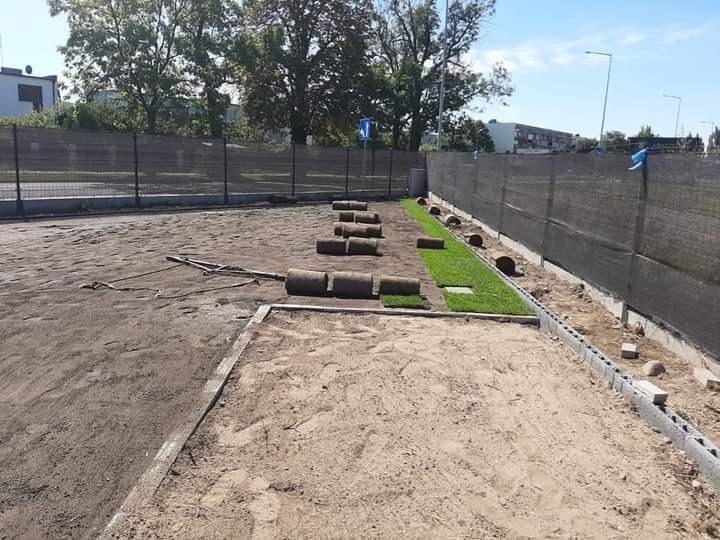 Rozwinięte i zwinięte rolki trawy na przygotowanym gruncie, widoczne ogrodzenie i budynek w tle, słoneczny dzień.