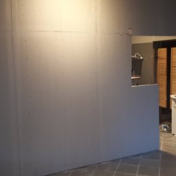 Usługi Elewacyjne Budowlane - Montaż płyt gipsowo-kartonowych na ścianie w trakcie remontu łazienki, widoczny otwór do pomieszczenia z umywalką i brązowymi żaluzjami.