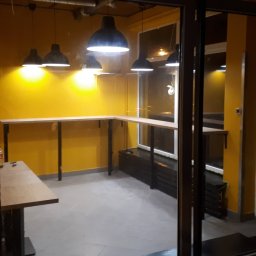 Usługi Elewacyjne Budowlane - Wnętrze lokalu z żółtymi ścianami i zamontowanymi drewnianymi blatami, widoczne przez szklane drzwi. Nad blatami wiszą czarne lampy. Wnętrze w trakcie wykańczania, widoczne niedokończone elementy.