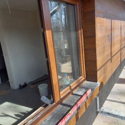 Usługi Elewacyjne Budowlane - Elewacja budynku z drewnopodobnym wykończeniem, widoczne okno z otwartą ramą, poziomica na parapecie, wiadro i rusztowanie na zewnątrz, surowe wnętrze w tle.