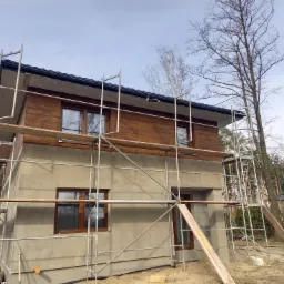 Dwukondygnacyjny dom w trakcie ocieplania elewacji, z widocznym rusztowaniem, częściowo otynkowaną ścianą i fragmentem drewnianej elewacji na górnej kondygnacji.