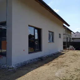 Elewacja domu jednorodzinnego po nałożeniu jasnego tynku, okna zabezpieczone folią, widoczne rusztowania i przygotowane farby.
