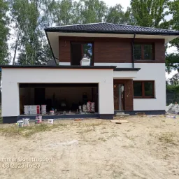 Dwukondygnacyjny dom z częściowo pomalowaną elewacją w kolorach bieli i brązu, widoczne puszki z farbą i drabina w garażu, teren wokół domu piaszczysty.