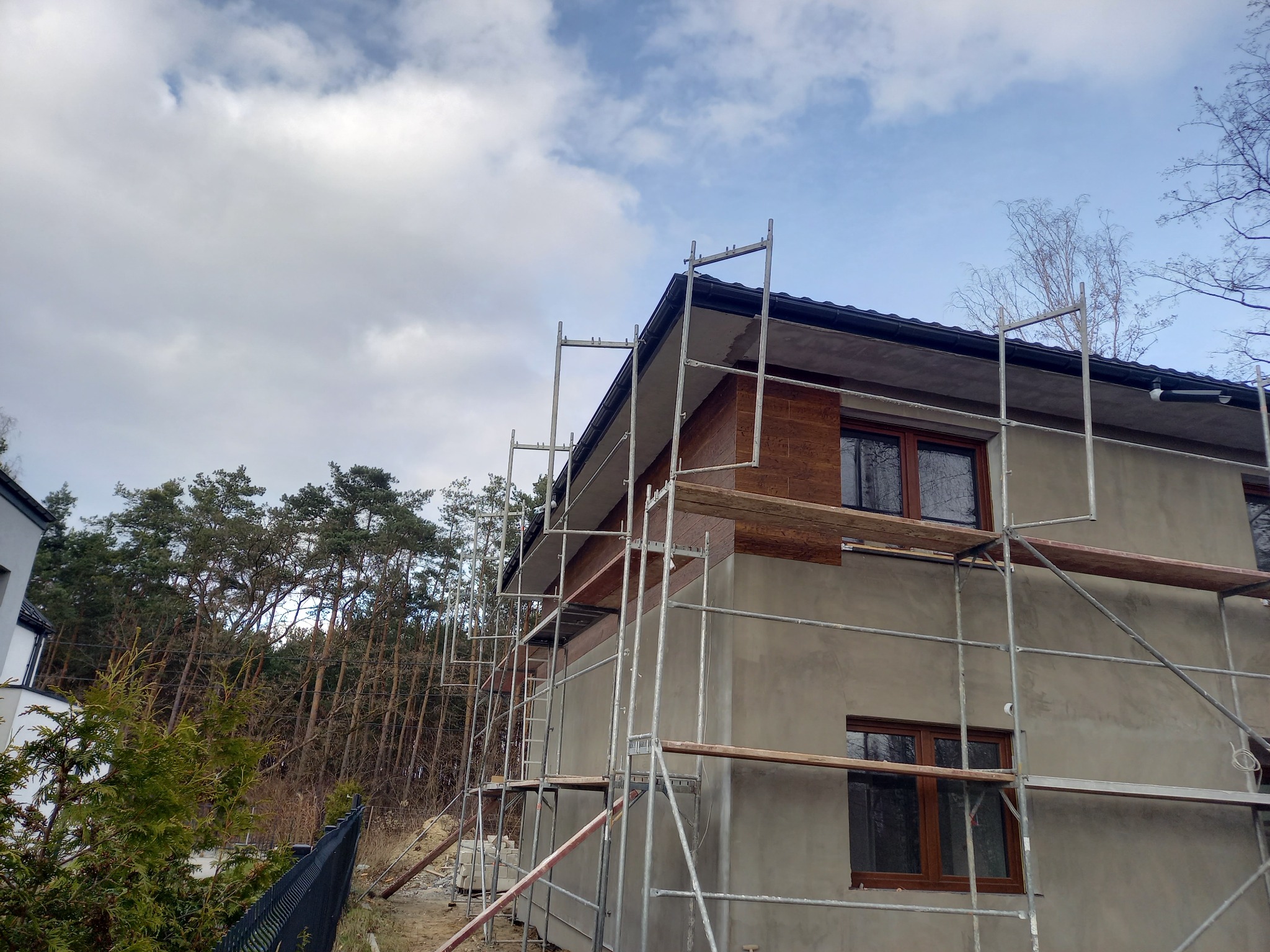 Dom jednorodzinny w trakcie ocieplania elewacji, widoczne rusztowania i fragment ocieplonej ściany z drewnopodobną okładziną, tło stanowią drzewa i pochmurne niebo.